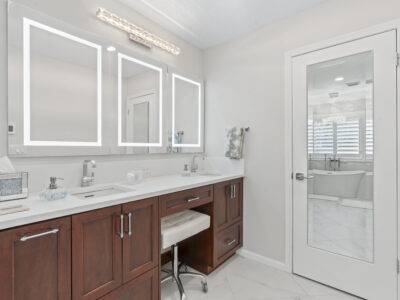 02-DSC04367 Modern bathroom with sleek white countertops, wooden cabinetry, illuminated mirrors, and a glass door leading to a bright space, emphasizing stylish design and functionality in home remodeling.