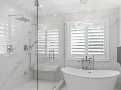 07-DSC04308 Modern bathroom featuring a freestanding bathtub, glass shower enclosure, marble walls, and natural light from window with shutters, highlighting elegant design elements for remodeling inspiration.