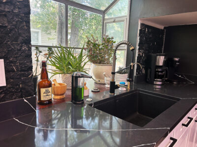 Chris1 Modern kitchen countertop with dark stone surface, sink, faucet, coffee maker, and plants, showcasing a stylish and functional design for kitchen remodeling.