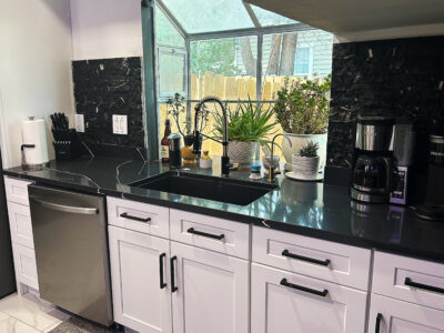 Chris2 Modern kitchen countertop featuring sleek black granite, white cabinetry, stainless steel dishwasher, and coffee maker, surrounded by decorative plants and natural light.