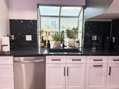 Chris3 Modern kitchen with black countertop, stainless steel dishwasher, white cabinetry, and plants near window, showcasing a stylish remodeling design.