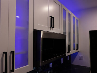 Chris8 Modern kitchen cabinetry with glass panels, illuminated by blue lighting, featuring a stainless steel microwave, highlighting contemporary design elements for kitchen remodeling.
